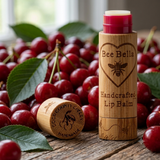Bee Bella handcrafted lip balm in a bamboo tube with a cherry-colored balm visible at the top, shown with the cap off beside fresh cherries and green leaves on a rustic wooden surface.