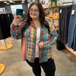 A woman takes a mirror selfie in a clothing store wearing a colorful plaid button-up shirt over a white top and black pants.