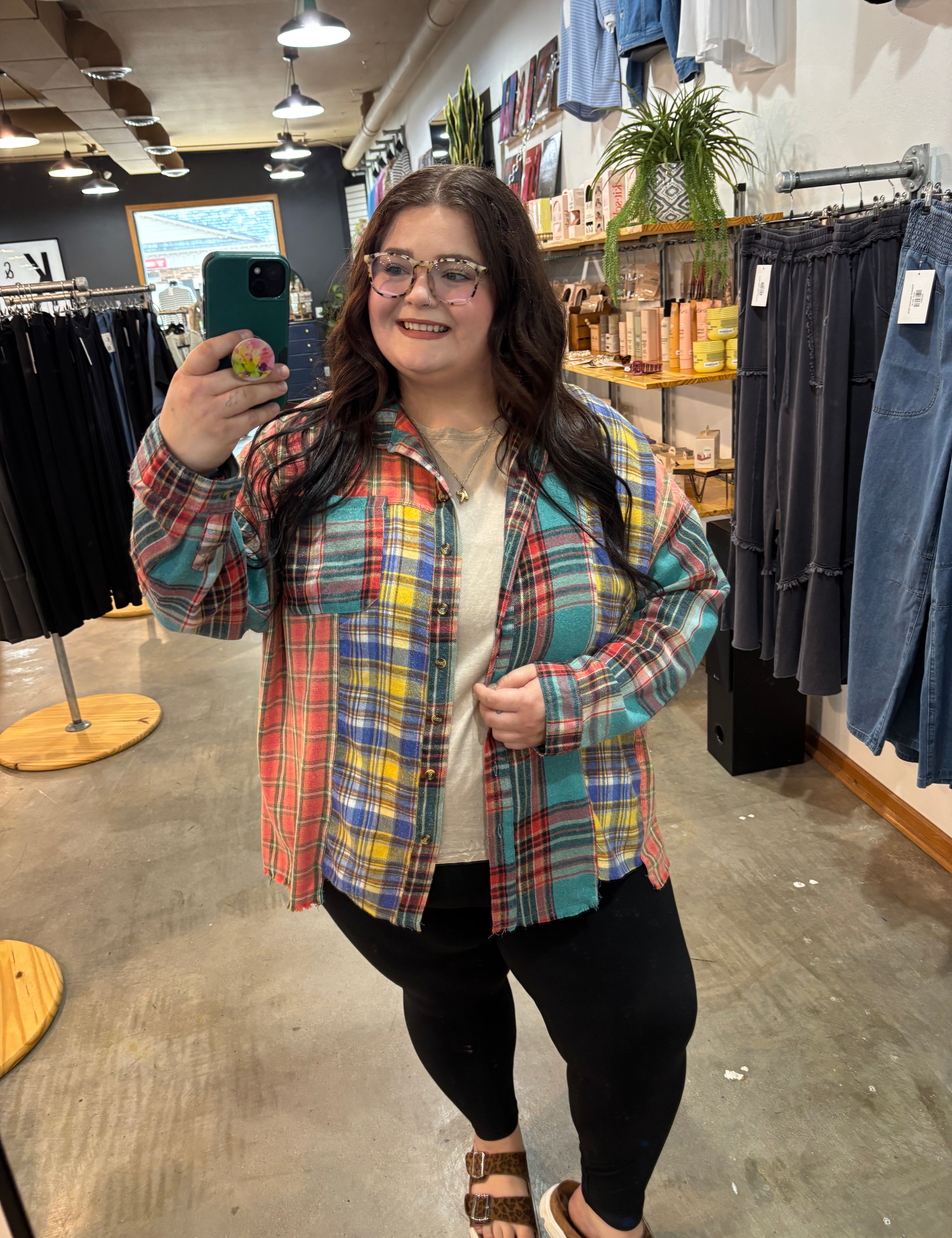 A woman takes a mirror selfie in a clothing store wearing a colorful plaid button-up shirt over a white top and black pants.