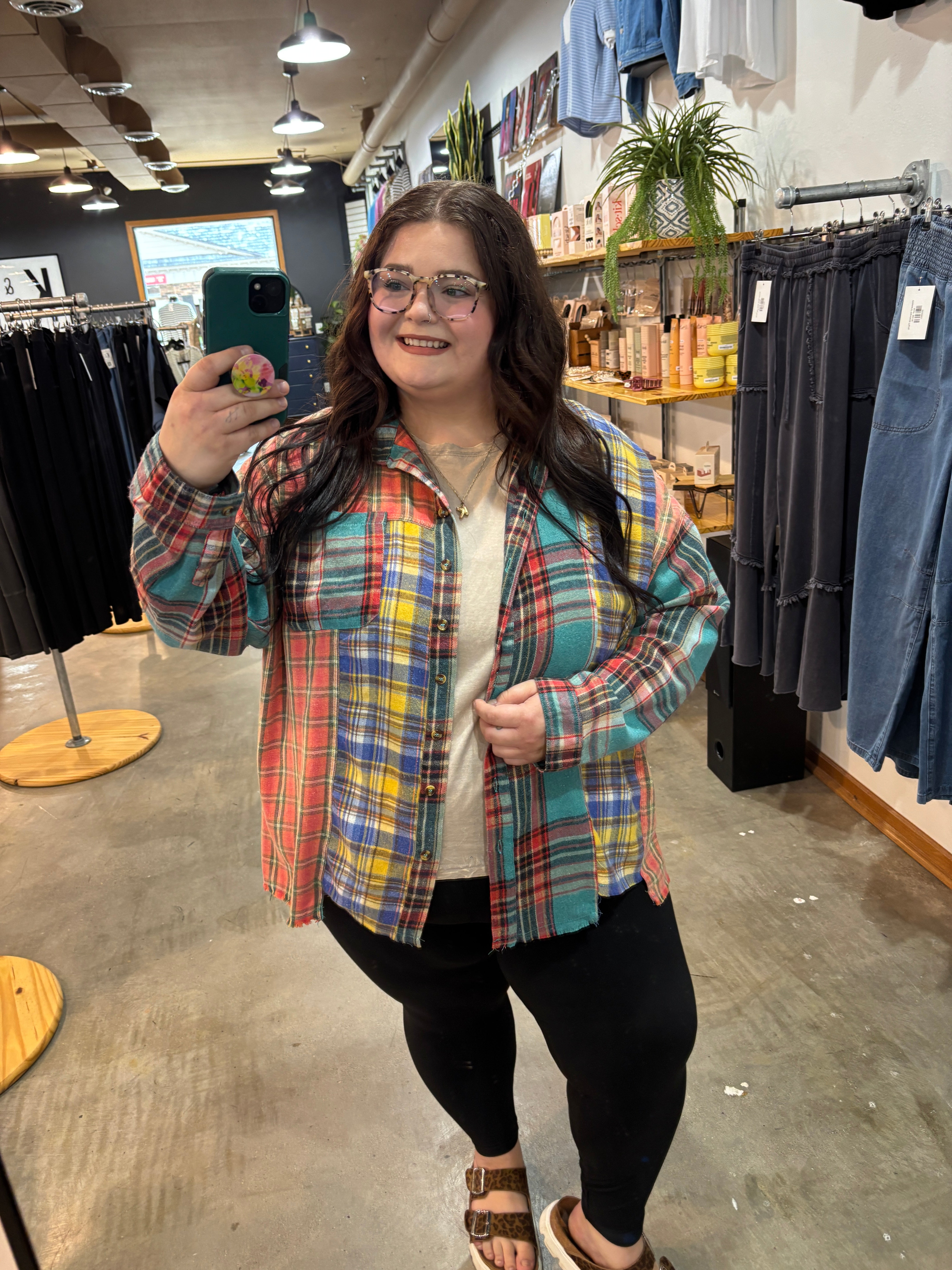 A woman takes a mirror selfie in a clothing store wearing a colorful plaid button-up shirt over a white top and black pants.