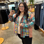 A woman taking a mirror selfie in a clothing store, wearing a colorful plaid button-up shirt over a cream t-shirt and black pants.