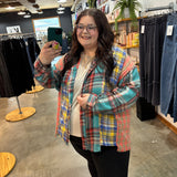 A woman taking a mirror selfie in a clothing store, wearing a colorful plaid button-up shirt over a cream t-shirt and black pants.