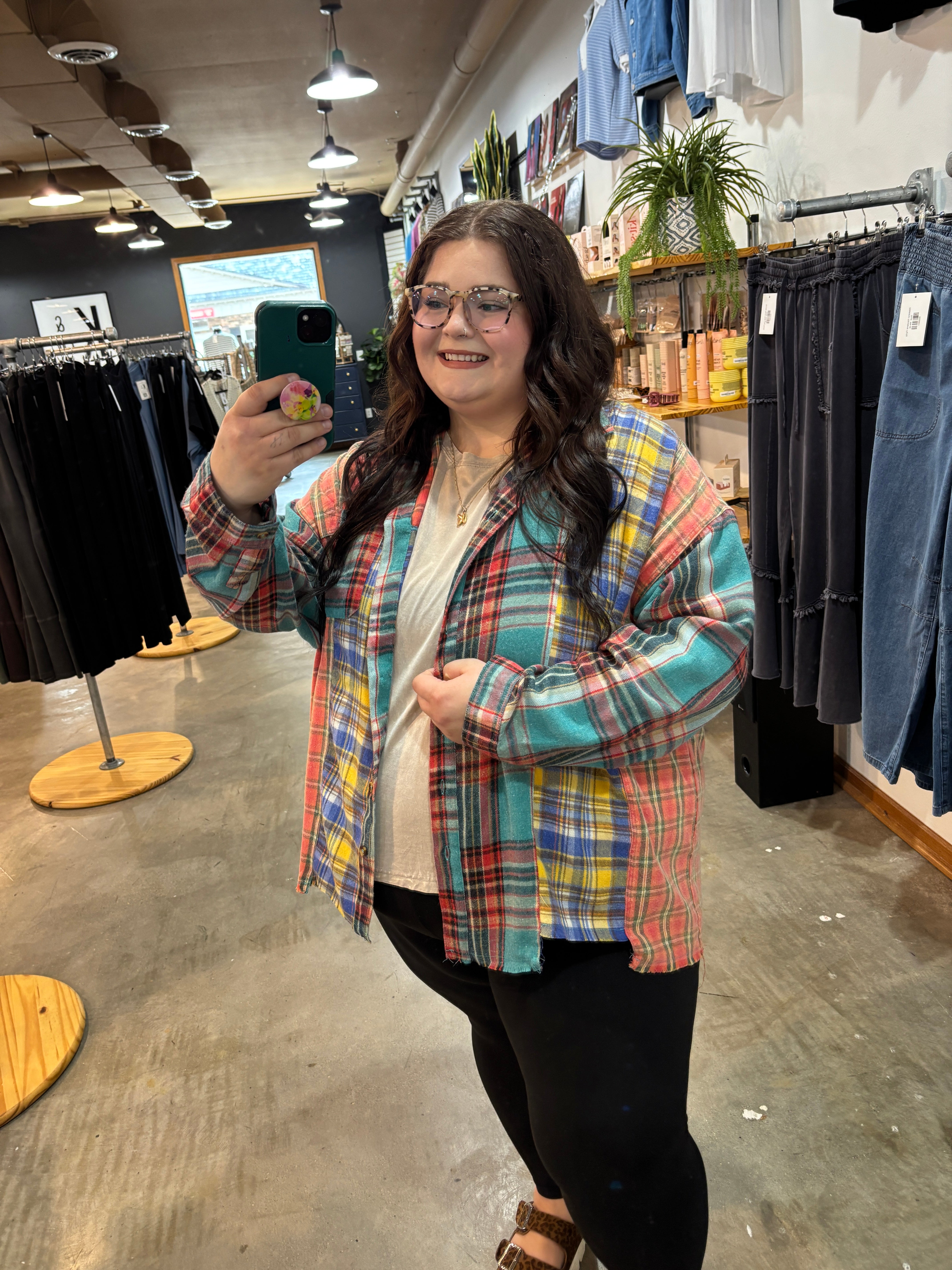 A woman taking a mirror selfie in a clothing store, wearing a colorful plaid button-up shirt over a cream t-shirt and black pants.