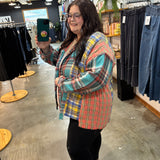 A woman taking a mirror selfie in a clothing store, wearing a colorful plaid button-up shirt with blue, red, yellow, and green tones, paired with black pants.