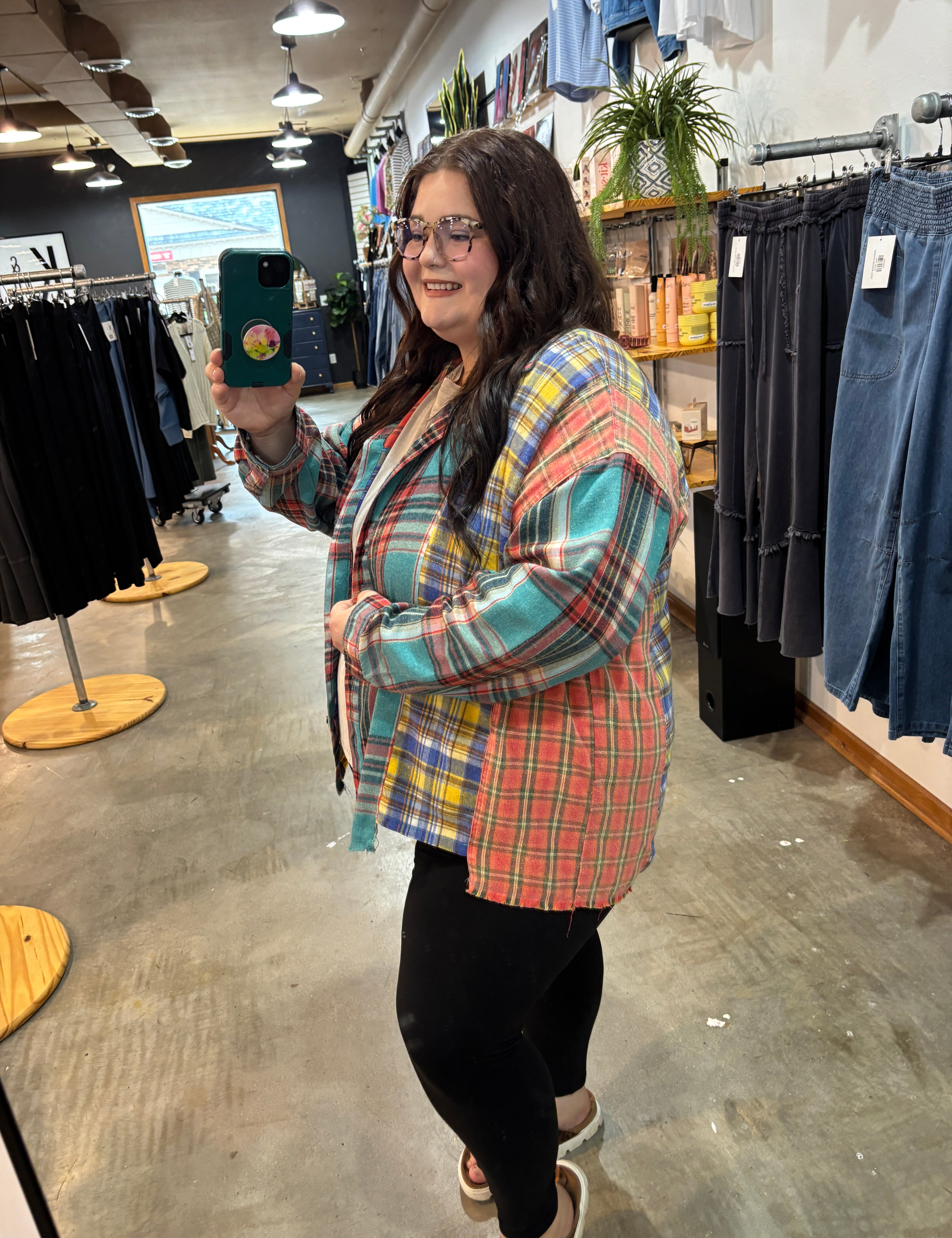 A woman taking a mirror selfie in a clothing store, wearing a colorful plaid button-up shirt with blue, red, yellow, and green tones, paired with black pants.