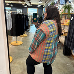 A woman takes a mirror selfie in a clothing boutique wearing an oversized plaid flannel shirt with red, blue, green, and yellow colors, paired with black leggings and brown sandals.