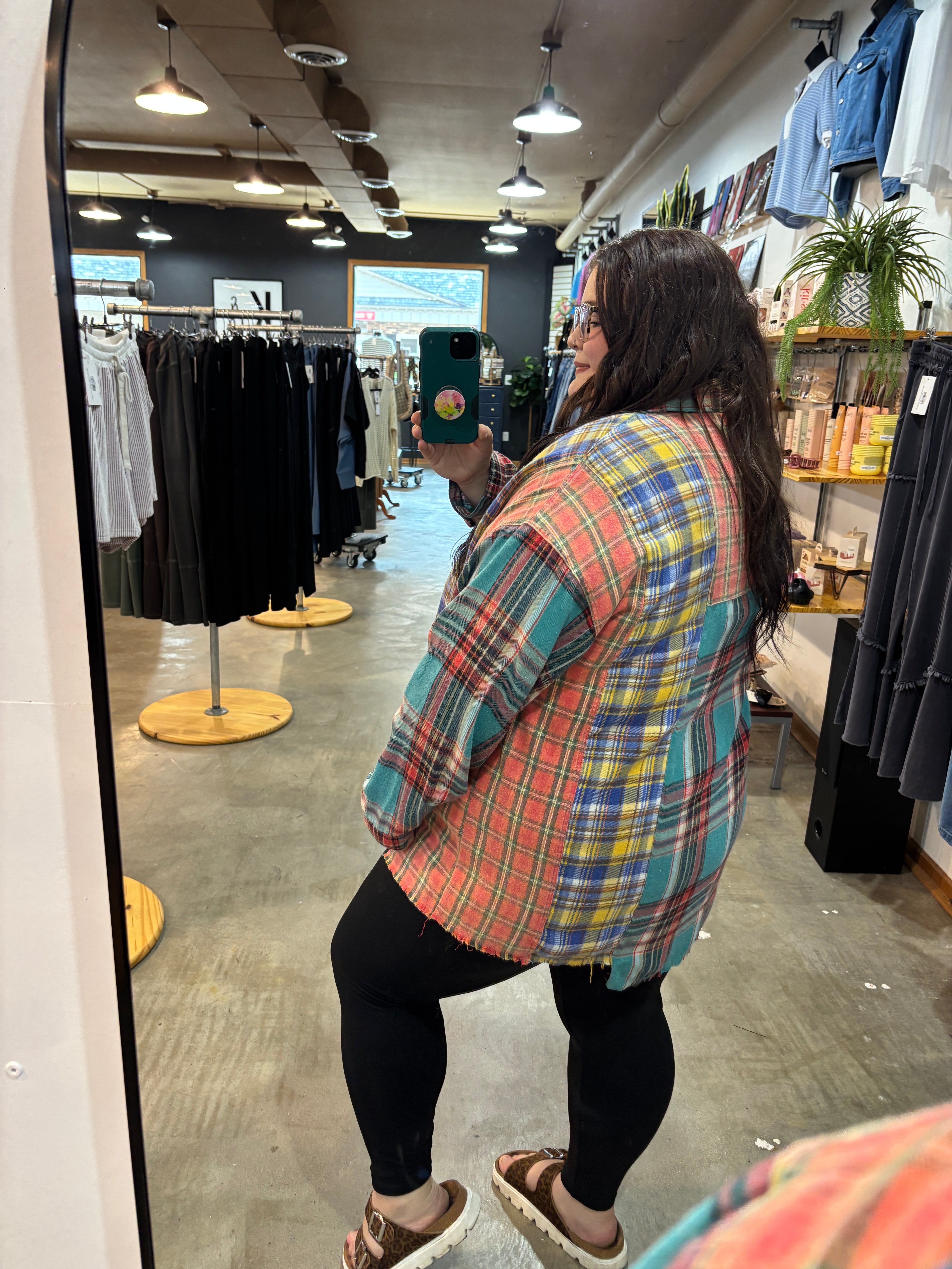 A woman takes a mirror selfie in a clothing boutique wearing an oversized plaid flannel shirt with red, blue, green, and yellow colors, paired with black leggings and brown sandals.