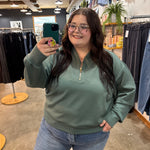 A woman taking a mirror selfie in a clothing store, wearing a sage green quarter-zip sweatshirt and light blue jeans.