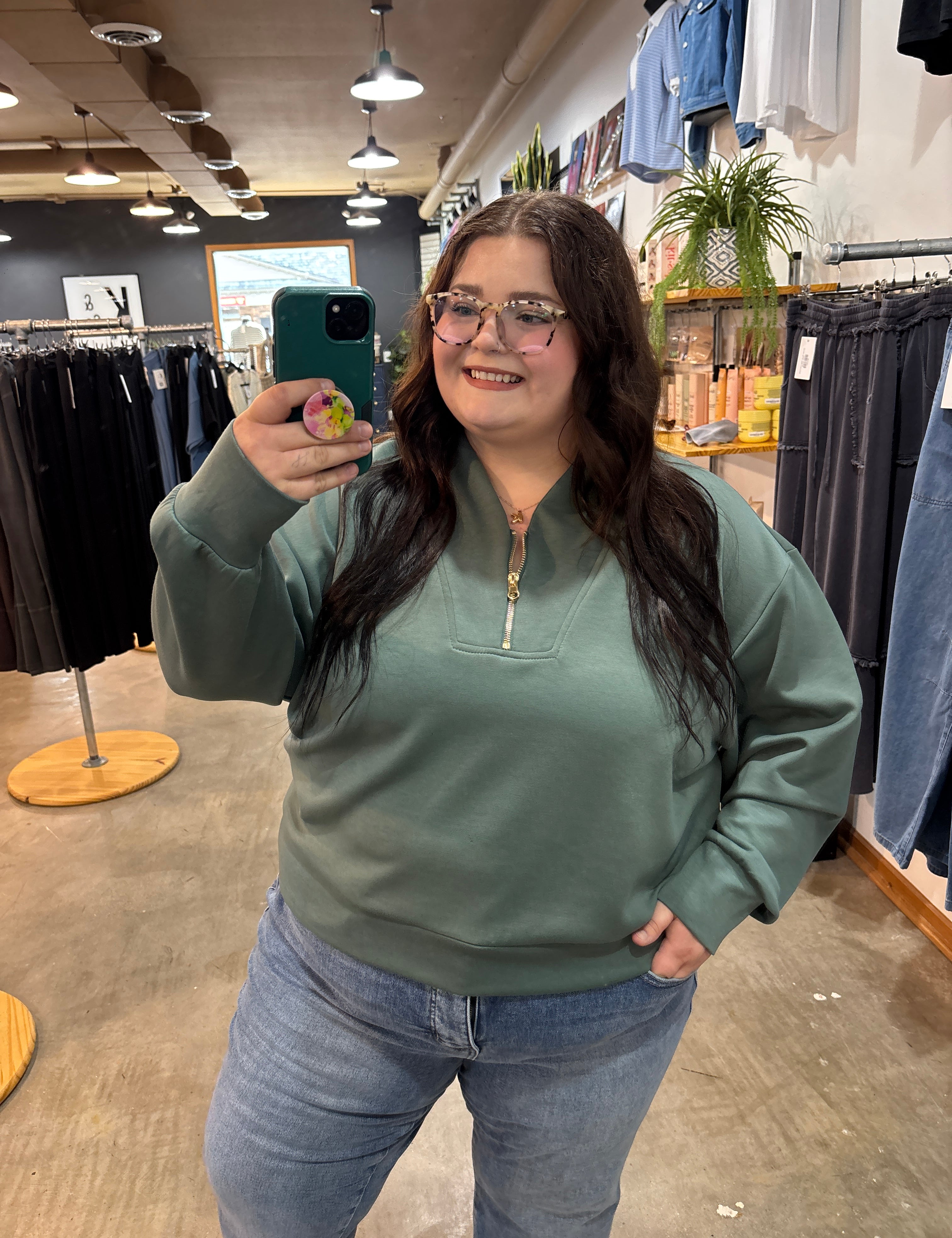 A woman taking a mirror selfie in a clothing store, wearing a sage green quarter-zip sweatshirt and light blue jeans.