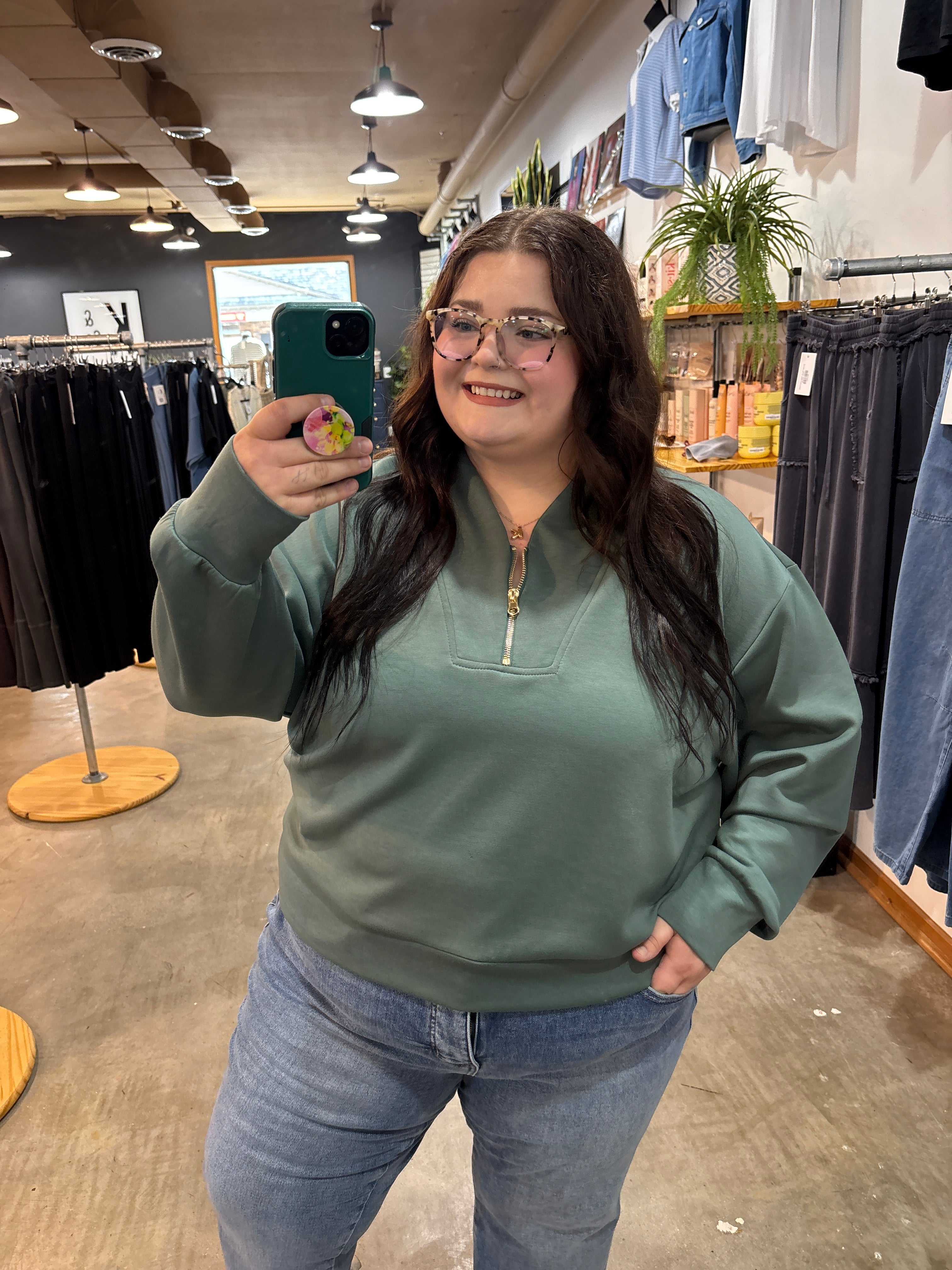 A woman taking a mirror selfie in a clothing store, wearing a sage green quarter-zip sweatshirt and light blue jeans.