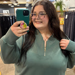 A woman taking a mirror selfie in a clothing store, wearing a sage green half-zip sweatshirt and glasses.