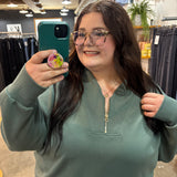 A woman taking a mirror selfie in a clothing store, wearing a sage green half-zip sweatshirt and glasses.