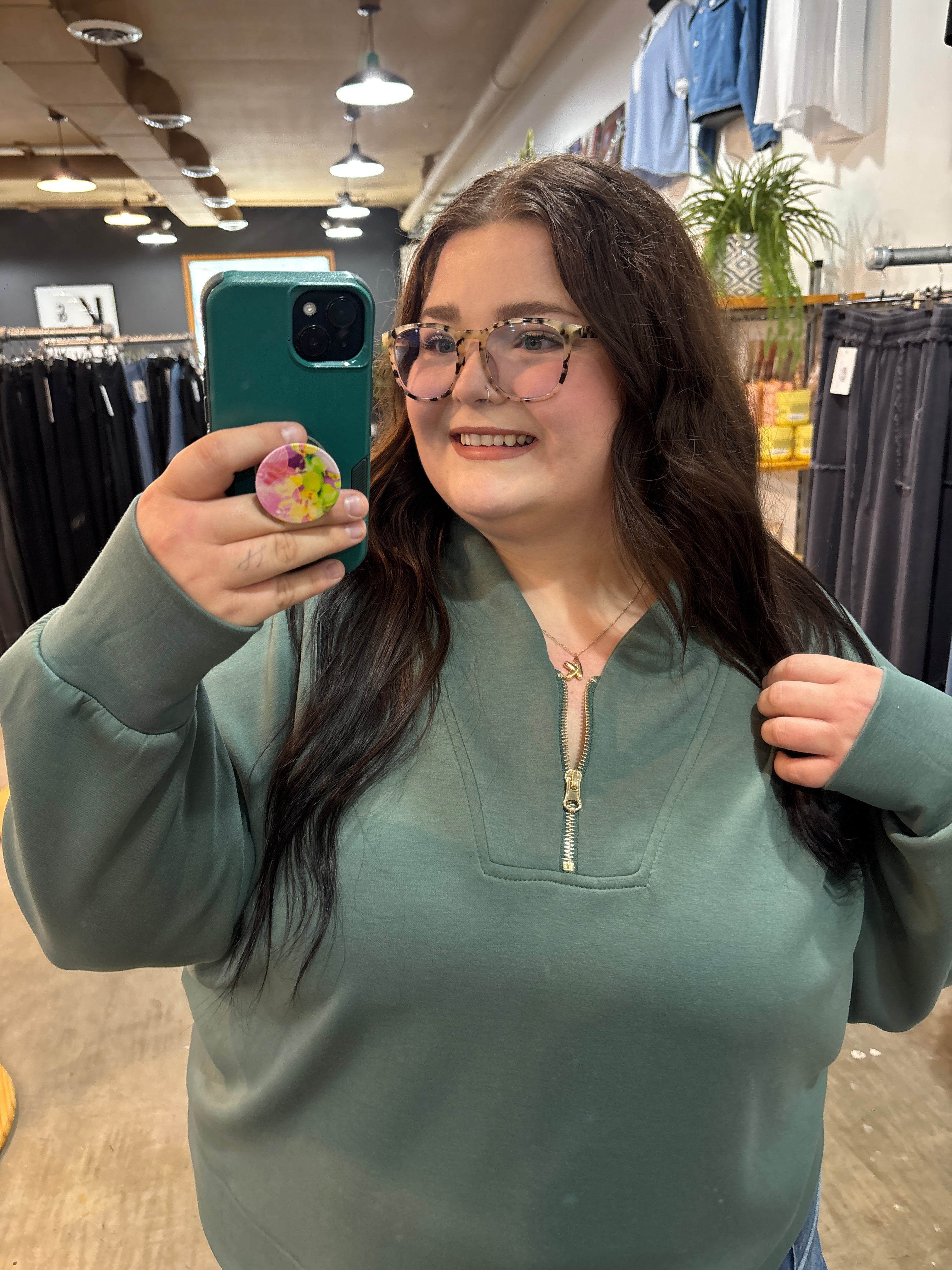 A woman taking a mirror selfie in a clothing store, wearing a sage green half-zip sweatshirt and glasses.