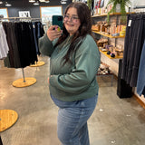 A woman taking a mirror selfie in a clothing store, wearing a sage green sweatshirt and blue jeans.