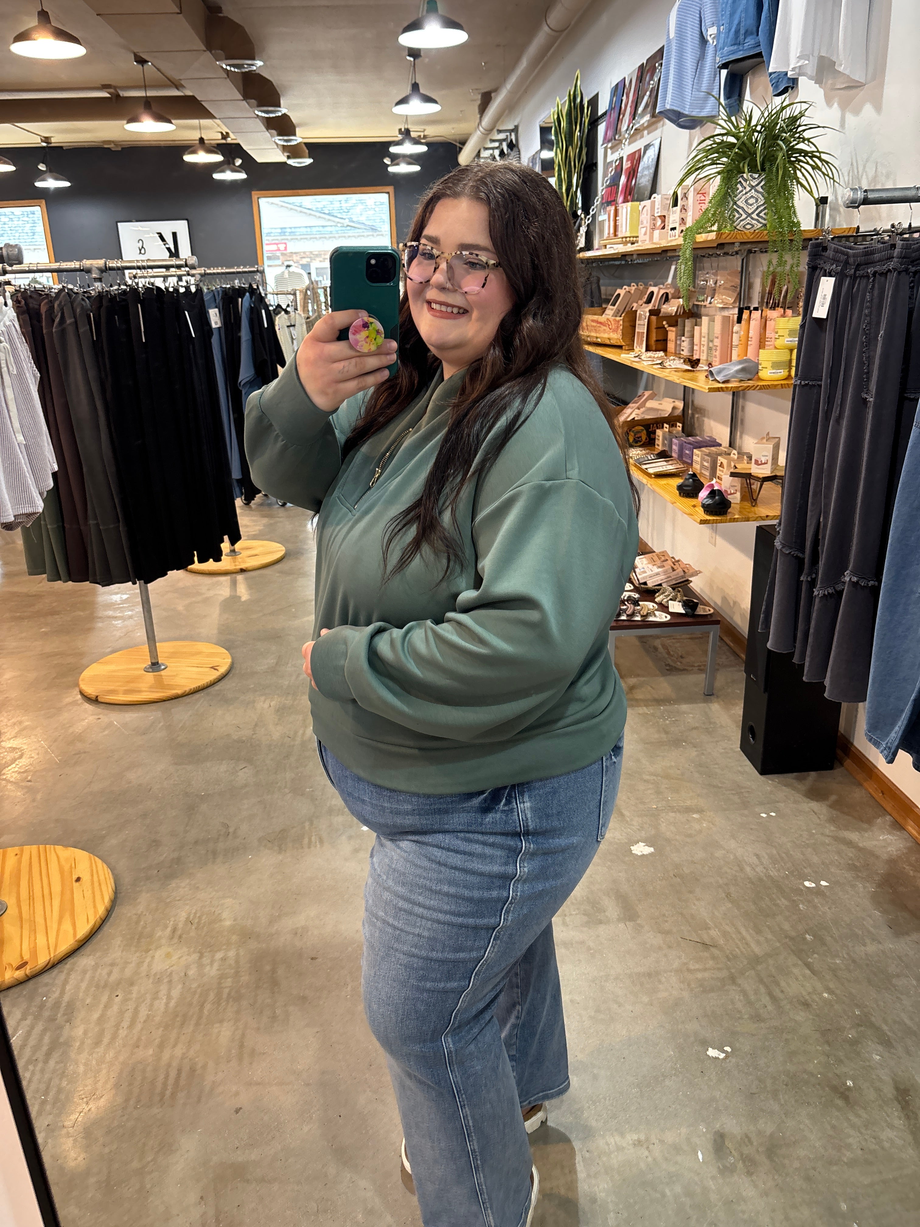 A woman taking a mirror selfie in a clothing store, wearing a sage green sweatshirt and blue jeans.