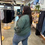 A woman taking a mirror selfie in a clothing store, wearing a sage green sweatshirt and light blue jeans.
