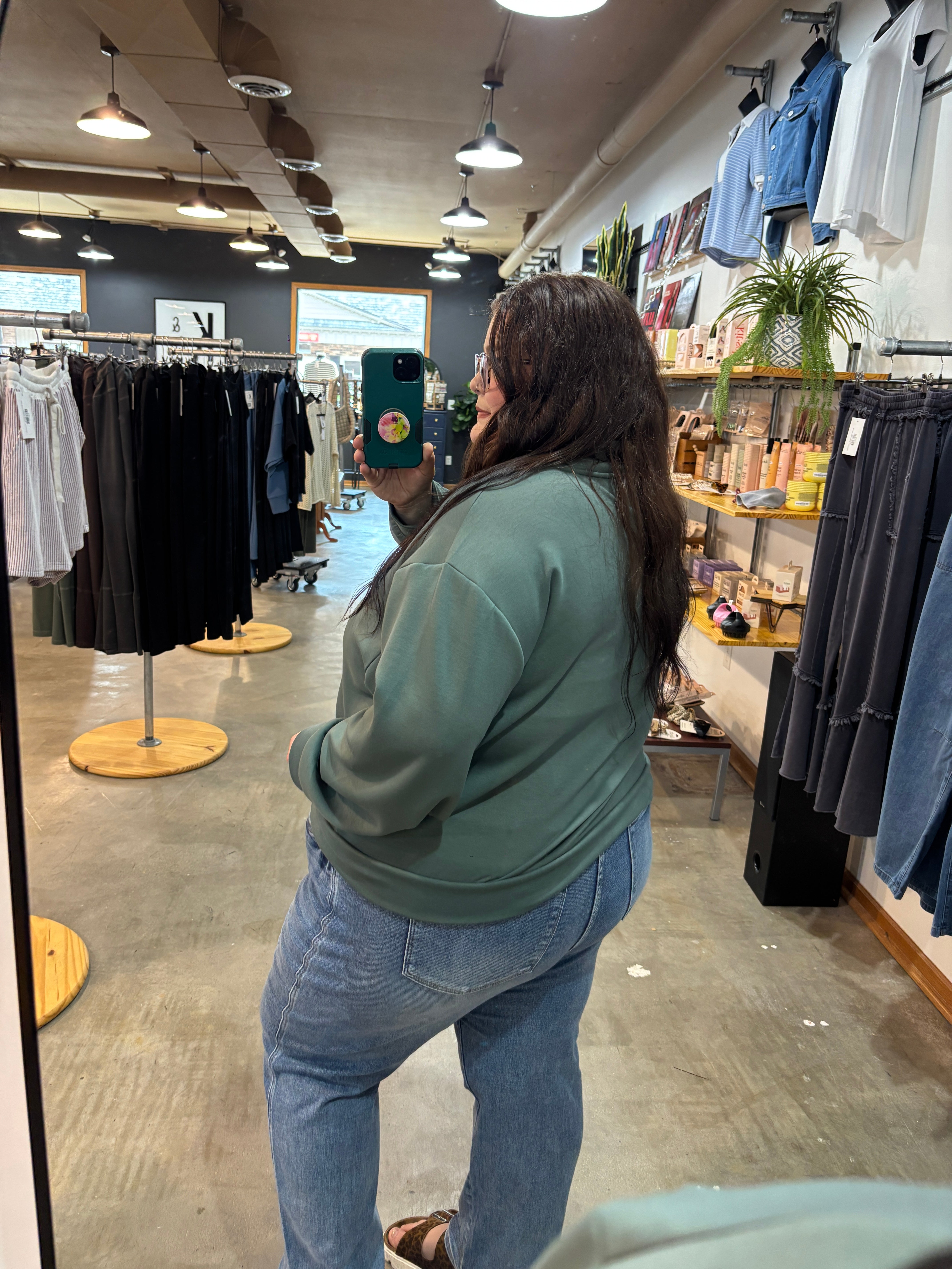 A woman taking a mirror selfie in a clothing store, wearing a sage green sweatshirt and light blue jeans.