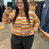 A woman taking a mirror selfie in a clothing store, wearing a colorful striped knit cardigan with yellow, orange, pink, and blue stripes, paired with dark gray corduroy pants.