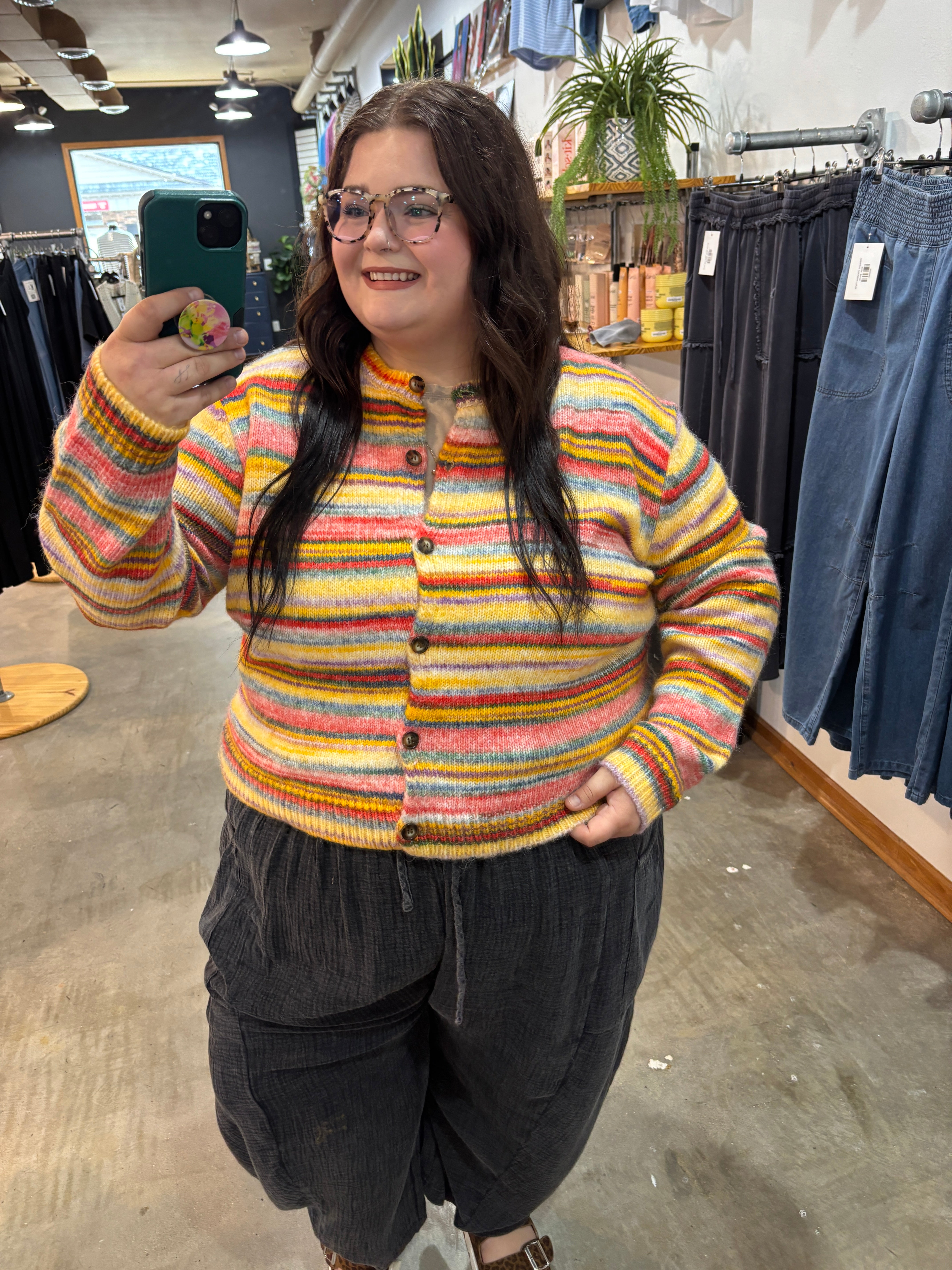 A woman taking a mirror selfie in a clothing store, wearing a colorful striped knit cardigan with yellow, orange, pink, and blue stripes, paired with dark gray corduroy pants.