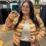 A woman taking a mirror selfie in a clothing store, wearing a colorful striped fuzzy knit cardigan over a cream top and dark pants.