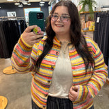 A woman taking a mirror selfie in a clothing store, wearing a colorful striped fuzzy knit cardigan over a cream top and dark pants.