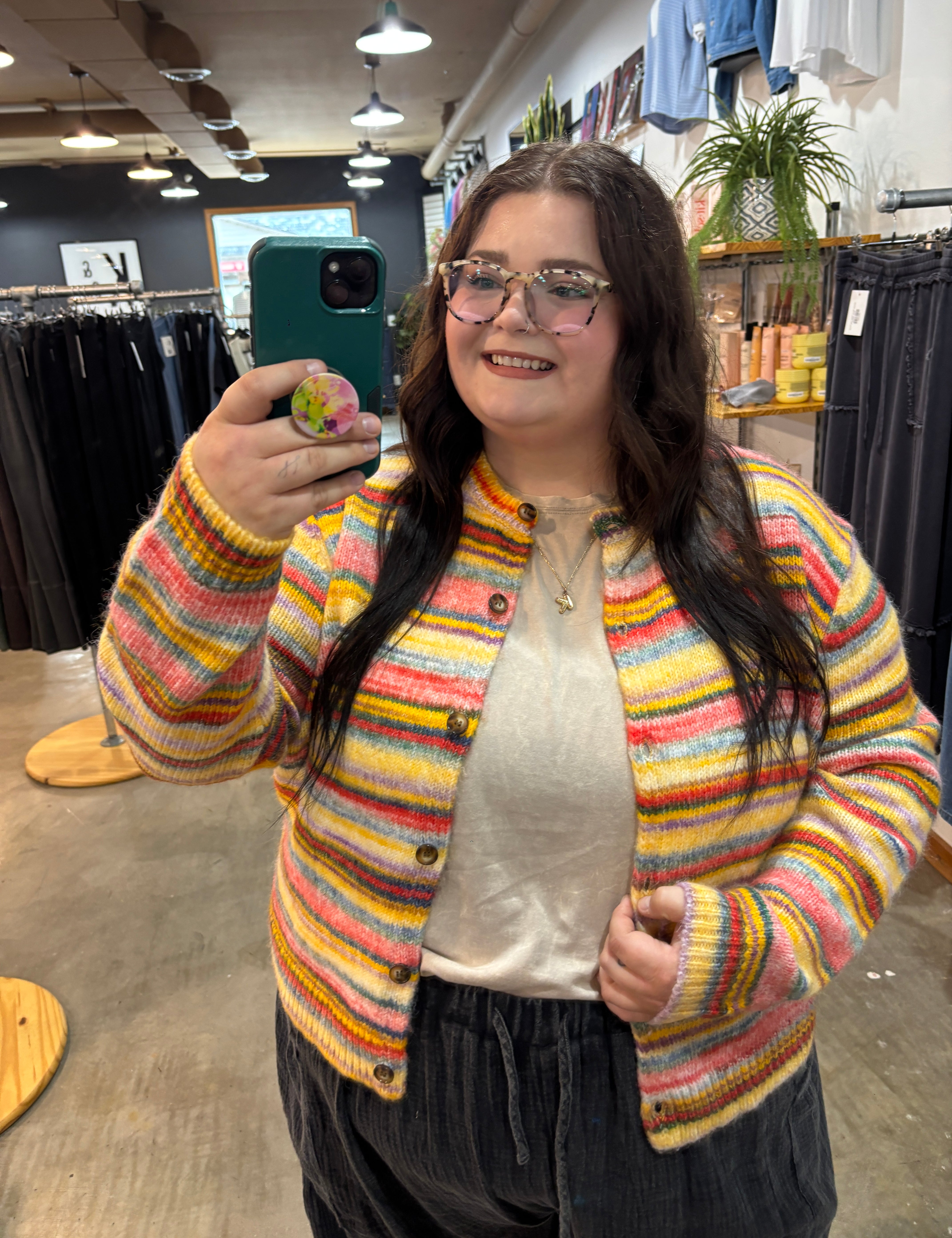 A woman taking a mirror selfie in a clothing store, wearing a colorful striped fuzzy knit cardigan over a cream top and dark pants.