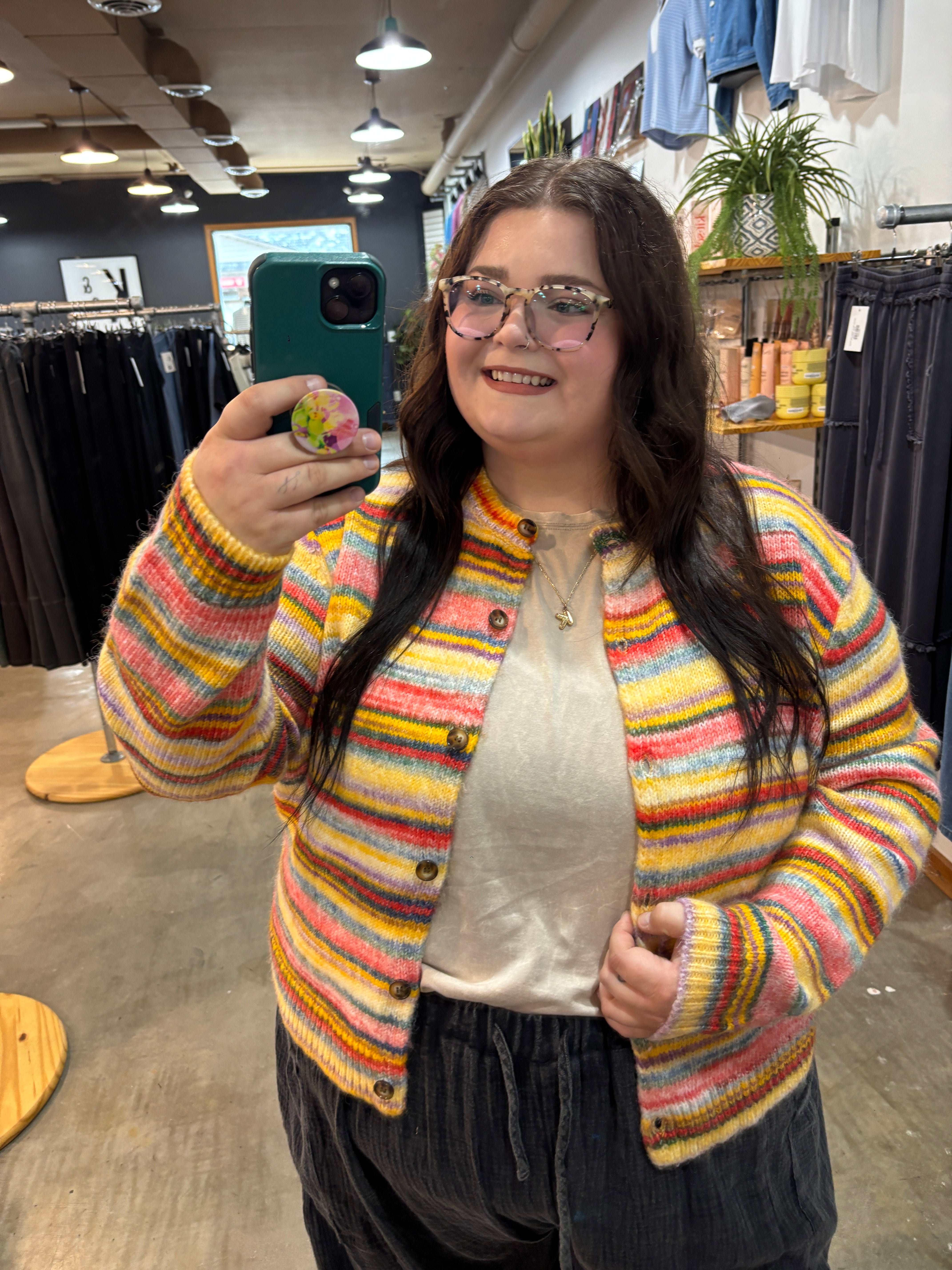 A woman taking a mirror selfie in a clothing store, wearing a colorful striped fuzzy knit cardigan over a cream top and dark pants.