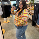 A woman taking a mirror selfie in a clothing boutique, wearing a colorful striped fuzzy knit sweater and blue jeans.