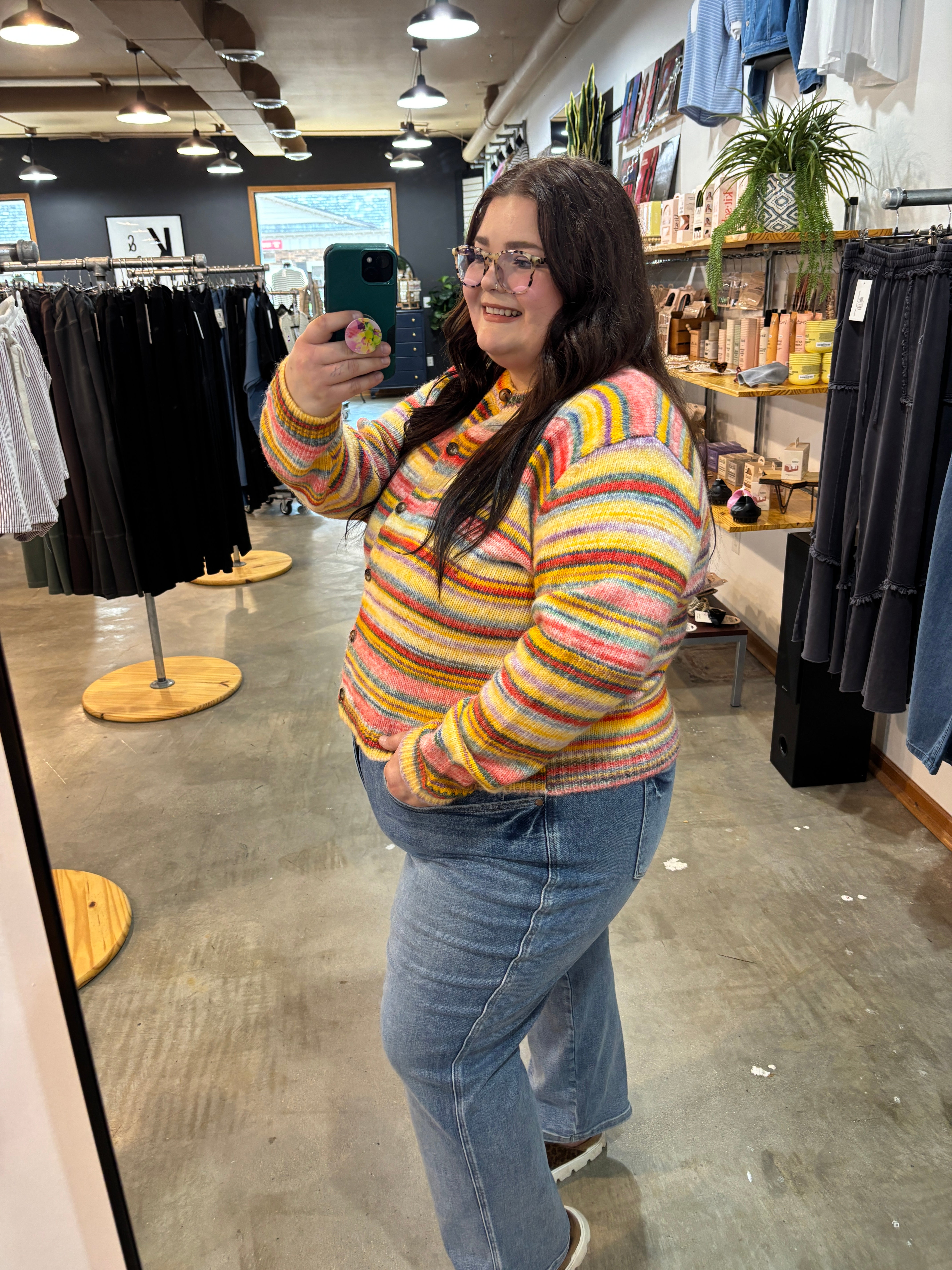 A woman taking a mirror selfie in a clothing boutique, wearing a colorful striped fuzzy knit sweater and blue jeans.