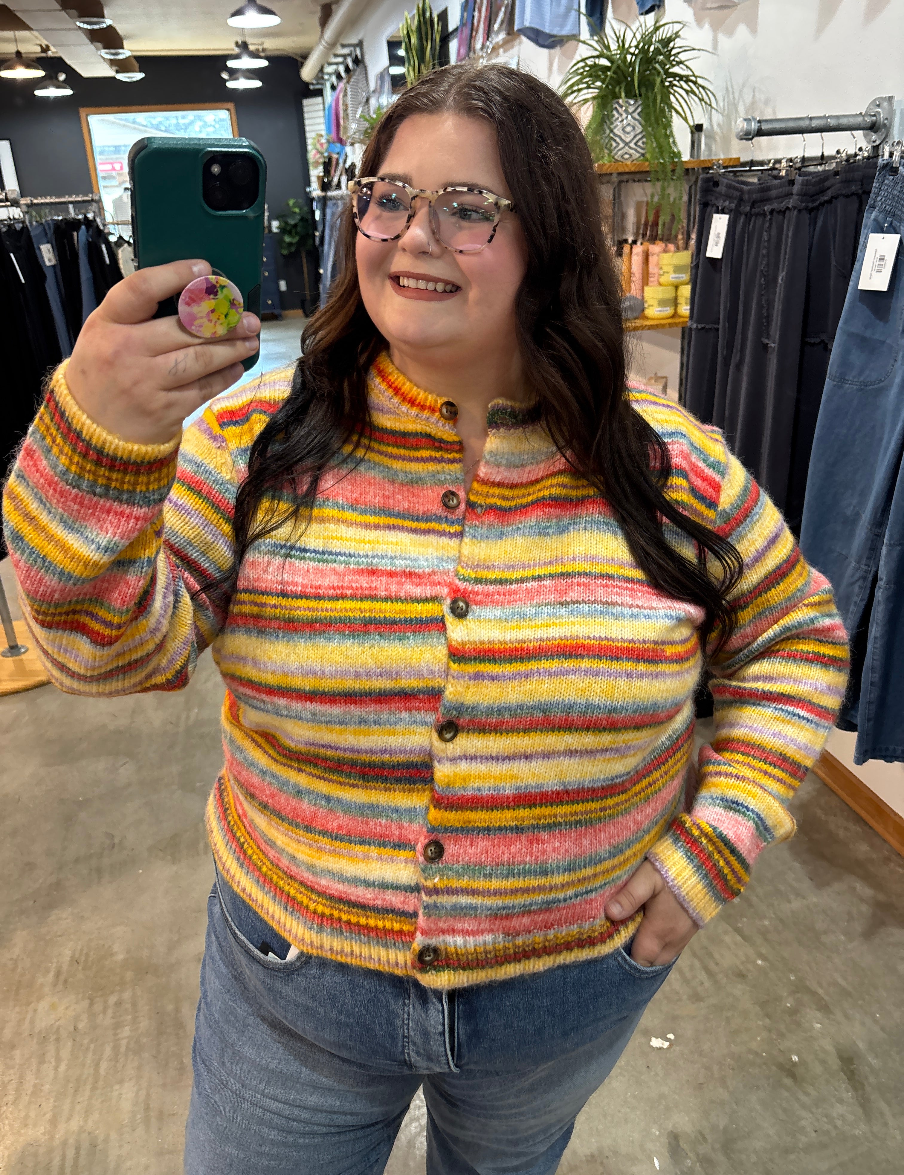 A woman taking a mirror selfie in a clothing store, wearing a colorful striped knit cardigan sweater with blue jeans.