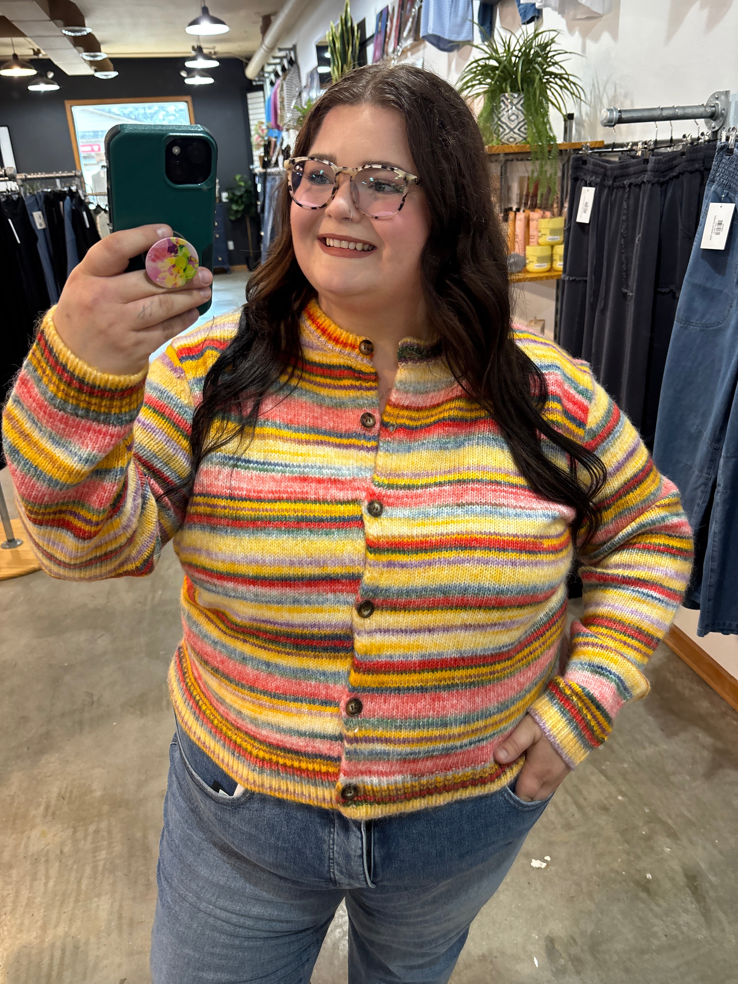 A woman taking a mirror selfie in a clothing store, wearing a colorful striped knit cardigan sweater with blue jeans.
