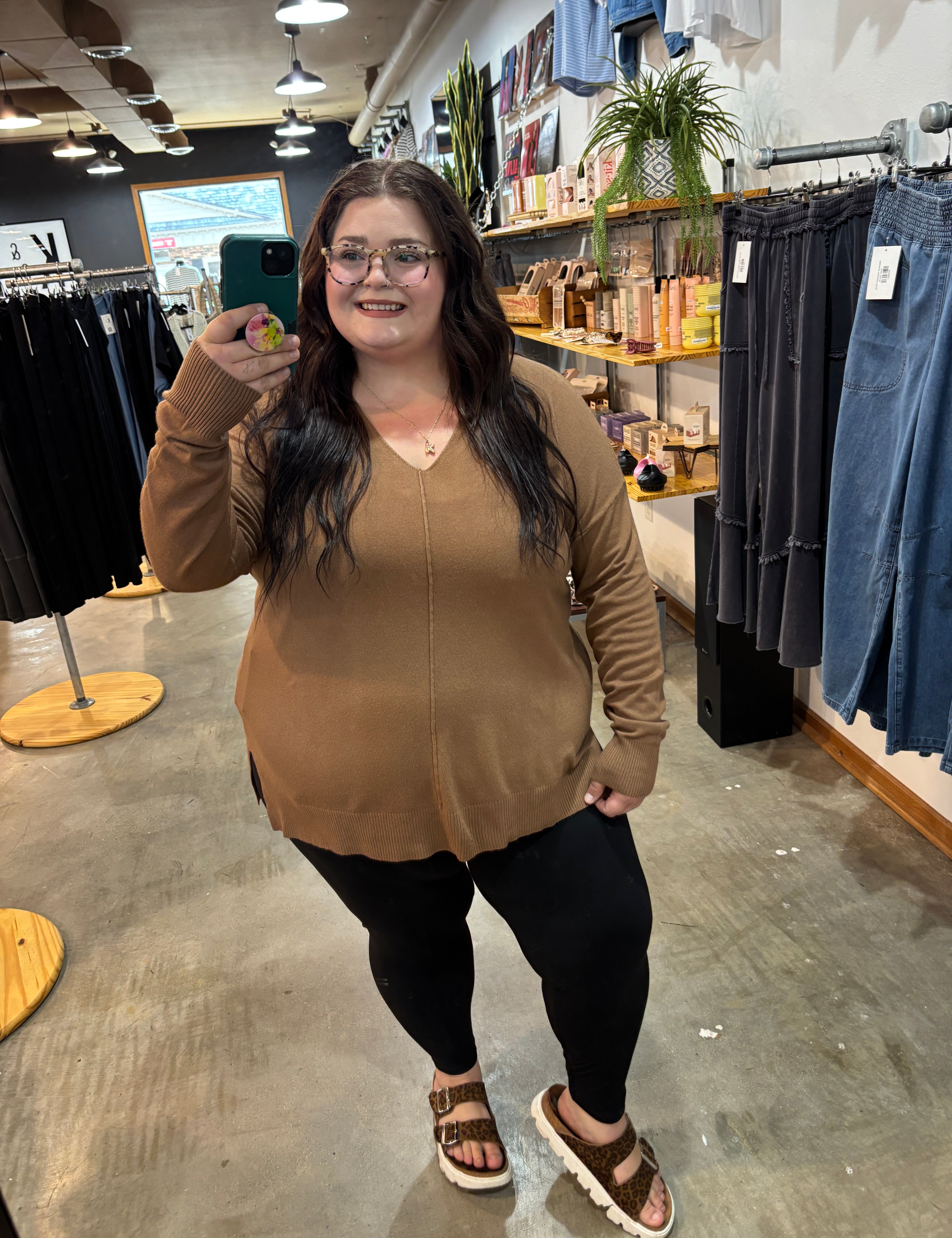 A woman takes a mirror selfie in a clothing store, wearing a tan long-sleeve knit sweater, black leggings, and brown sandals.