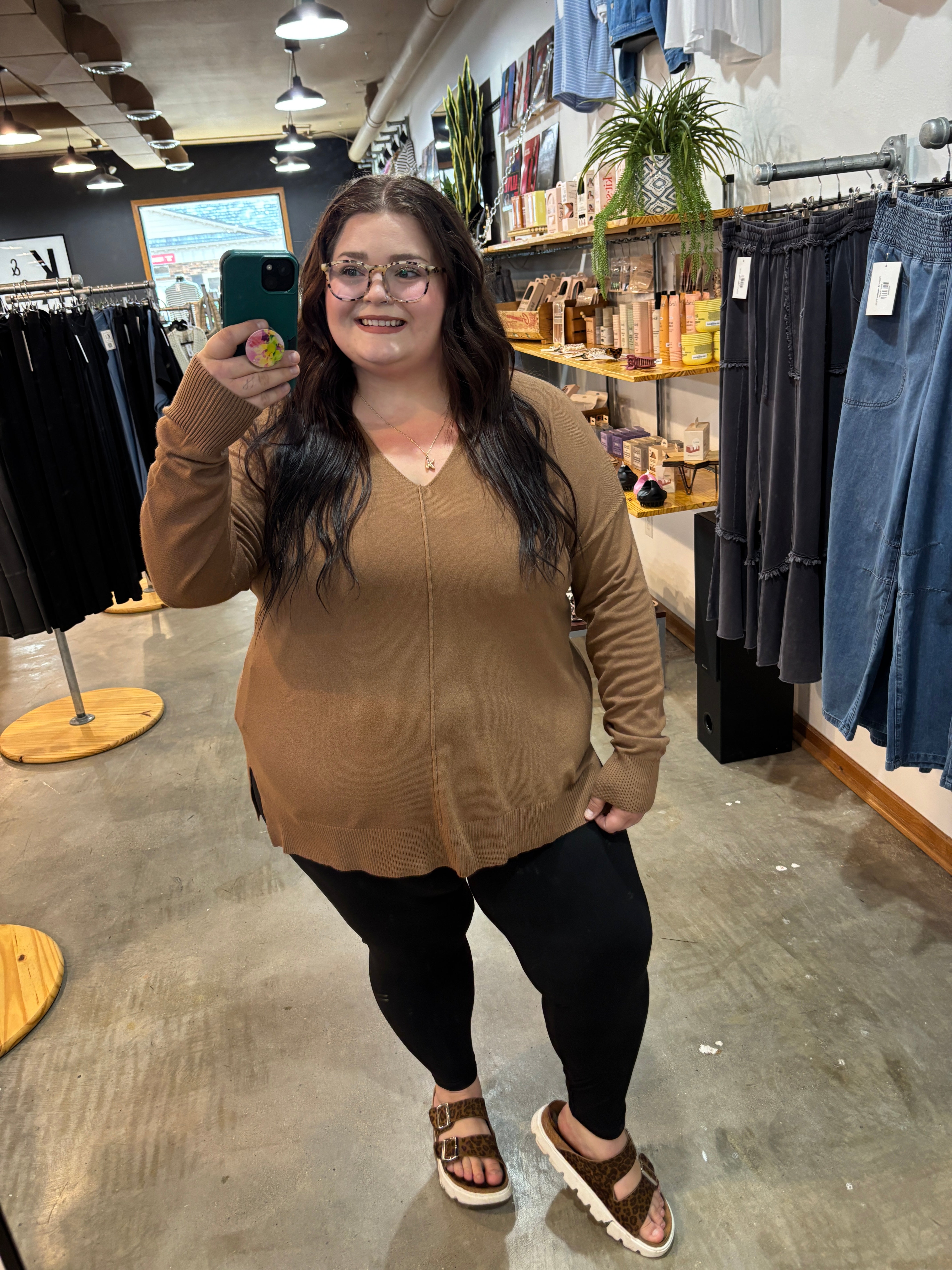 A woman takes a mirror selfie in a clothing store, wearing a tan long-sleeve knit sweater, black leggings, and brown sandals.